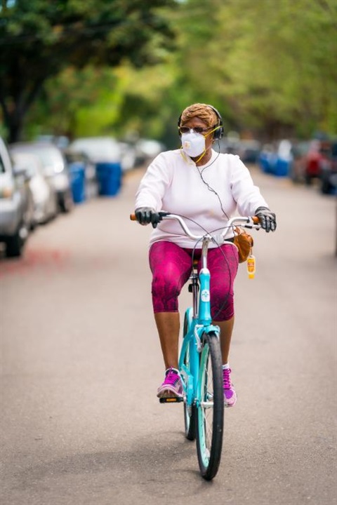 Woman with mask rides cruiser bike on a residential street. While plenty of parked cars are visible, there are no moving cars.